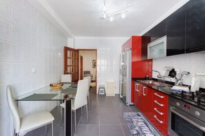 A modern kitchen with a glass dining table, white chairs, a red and black refrigerator, and a stainless steel oven.