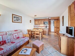 A living room with a patterned sofa, a wooden dining table, and a kitchen area with wooden cabinets and a tiled floor. - Ground floor, swimming pool, balcony, tv, ski locker, 50m², Les Menuires (Les Menuires)