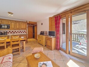 Eine Küche mit Holzausstattung, einem Esstisch und Stühlen. Ein Fernseher und ein kleines Schränkchen sind ebenfalls vorhanden. Eine Glastür öffnet sich zu einem Außenbereich mit einer Terrasse.