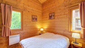 Un dormitorio de cabaña de madera con una cama, dos ventanas, un calentador y una lámpara.