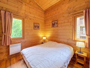 A wooden cabin bedroom with a bed, two windows, a heater, and a lamp. - 3rd floor, swimming pool, balcony, tv, ski locker, 50m², Les Menuires (Les Menuires)