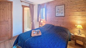 A bedroom with a wooden wall, a bed with a blue polka dot bedspread, a bedside table with a lamp, and a window with curtains. - 100m from ski slopes, 2nd floor, swimming pool, balcony, tv, ski locker, 46m², Les Menuires (Les Menuires)
