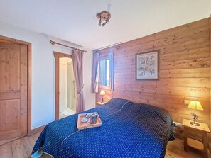 A bedroom with a wooden wall, a bed with a blue polka dot bedspread, a bedside table with a lamp, and a window with curtains. - 100m from ski slopes, 2nd floor, swimming pool, balcony, tv, ski locker, 46m², Les Menuires (Les Menuires)