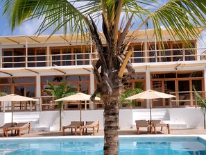 A pool area with lounge chairs and umbrellas, a palm tree, and a building with large windows and balconies. - Sunny villa matemwe (Matemwe)