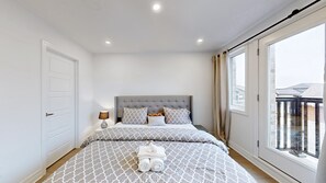 Ein Schlafzimmer mit grau gestepptem Kopfteil, gemusterten Betttextilien, einer Nachttischlampe und Blick auf einen Balkon durch ein großes Fenster.