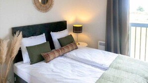 Een net opgemaakt bed met witte lakens en groene kussens, een bedlamp en een decoratieve spiegel aan de muur.