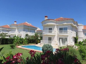 Un edificio grande de varios pisos con piscina, rodeado de jardines cuidados y plantas tropicales.