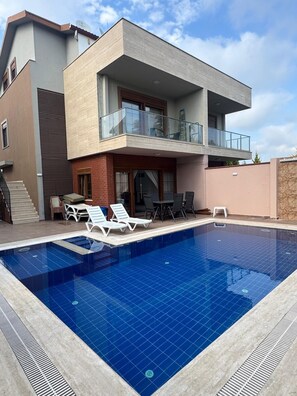 Una casa moderna de dos pisos con piscina, mobiliario de exterior y un balcón.