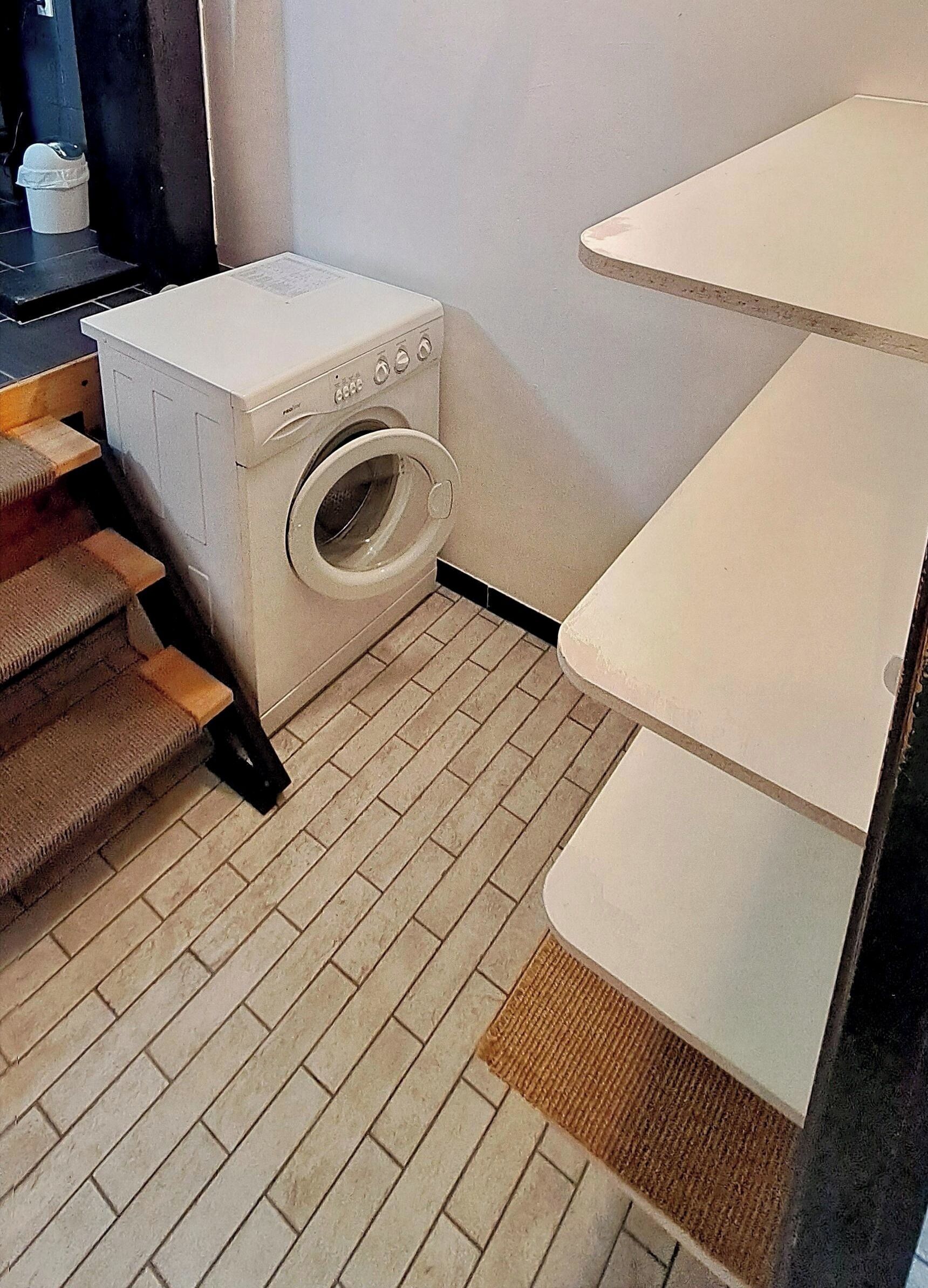 Een wasruimte met een wasmachine, een kleine prullenbak en een trap naar een hoger niveau.