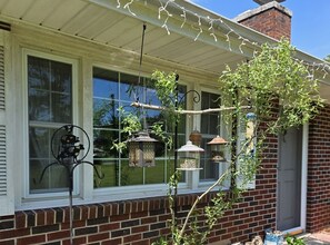 A house with a brick exterior, a white window with a fan, and hanging bird feeders. - Peaceful Home Near The Blue Ridge Mountains (Stuart)