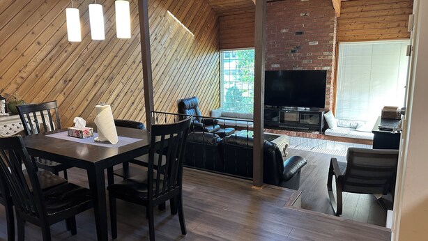 A dining area with a wooden table and chairs, a television in the background, and a brick wall.