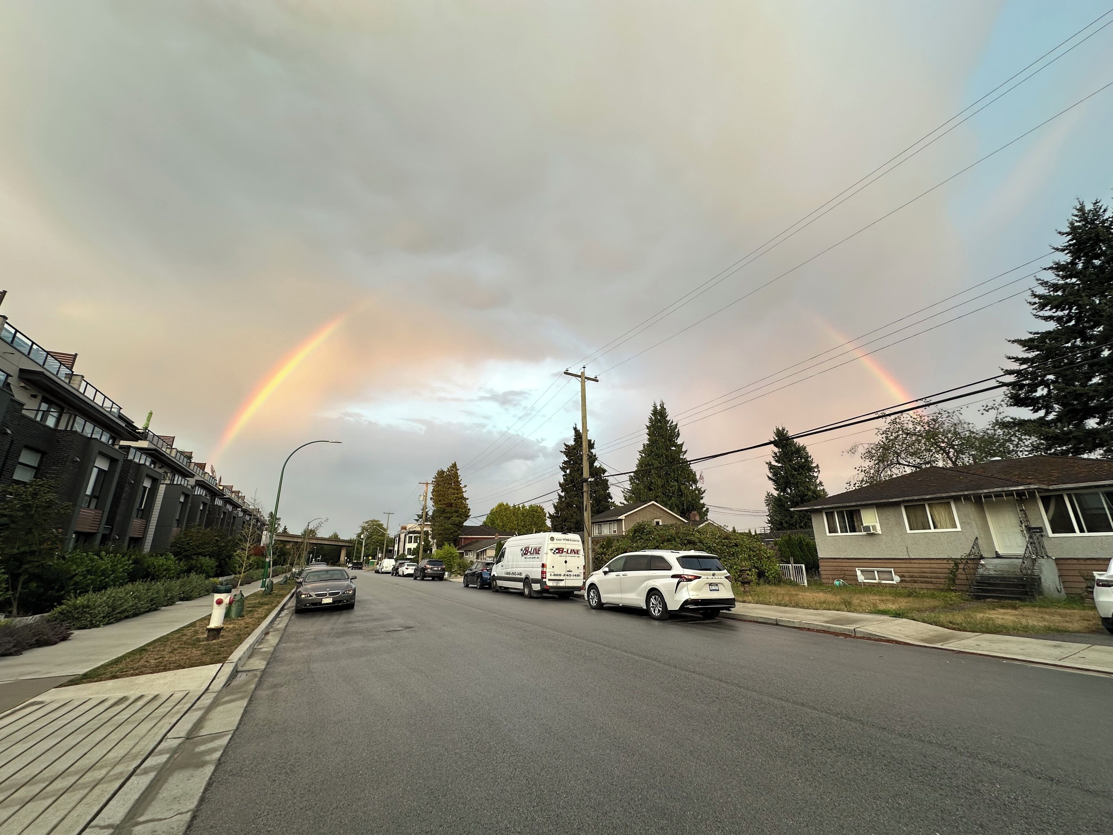 A residential street with houses, parked cars, and a rainbow in the sky.