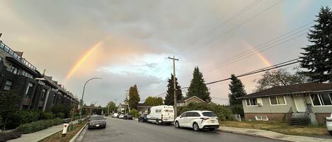A residential street with houses, parked cars, and a rainbow in the sky.