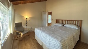 A bedroom with a wooden ceiling, a bed with patterned bedding, a chair, a floor lamp, and a window with blinds.