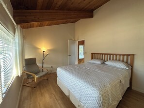 Ein Schlafzimmer mit Holzboden, einem Bett mit gemusterten Betttextilien, einem Sessel, einer Stehlampe und einem Fenster mit Jalousien.