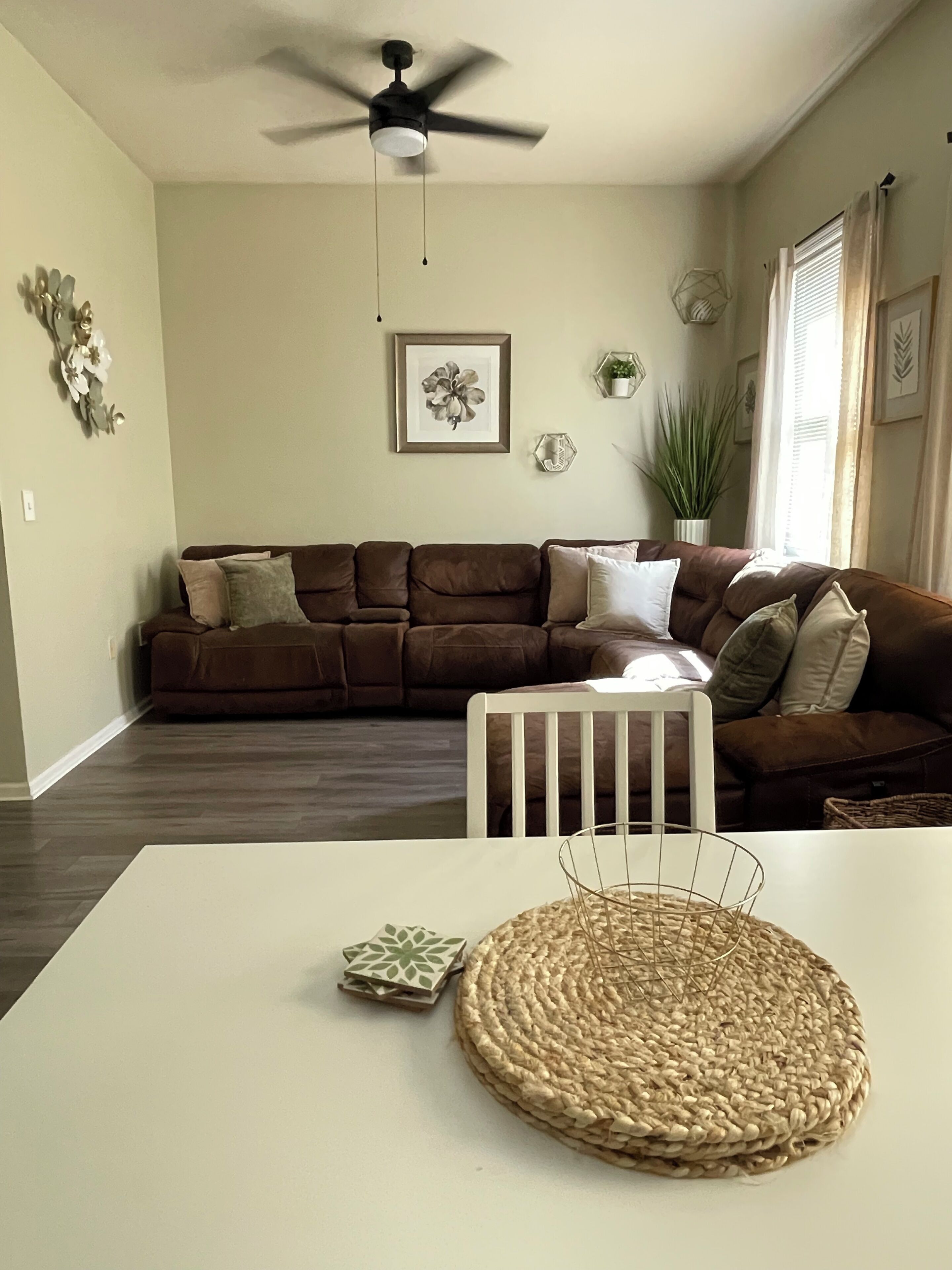 A living room with a brown sectional sofa, a white dining table, a ceiling fan, and a framed floral artwork on the wall.