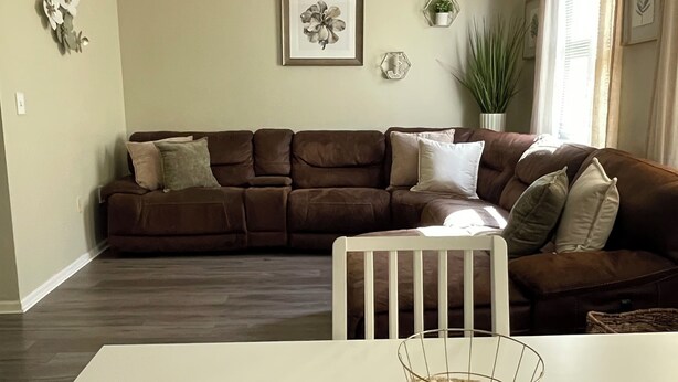 A living room with a brown sectional sofa, a white dining table, a ceiling fan, and a framed floral artwork on the wall. - Urban 2BR Near LightRail–Easy NYC & MetLife Access (Jersey City)