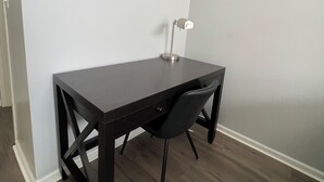 A corner room with a dark wooden desk, a black chair, a white wall, and a floor lamp. - Urban 2BR Near LightRail–Easy NYC & MetLife Access (Jersey City)