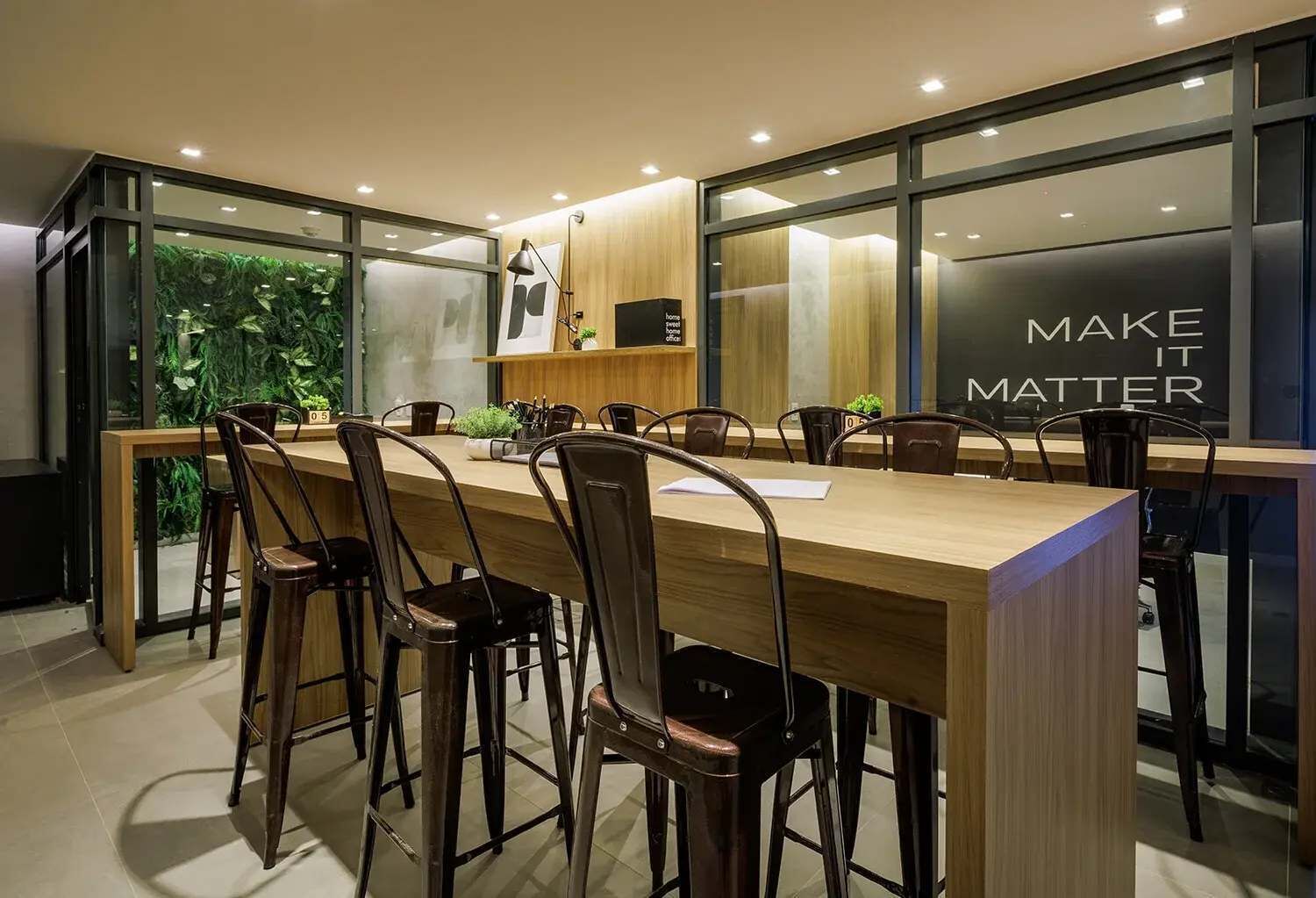 Un espace repas moderne avec une longue table en bois, de nombreuses chaises et un mur de verre orné d’une citation inspirante.