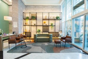 Un vestíbulo moderno con pared de ladrillo, sofá verde, sillas marrones y una mampara de vidrio con plantas.