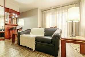 Et moderne hotelværelse med lædersofa, et træsidebord, et spejl og et stort vindue med gardiner.