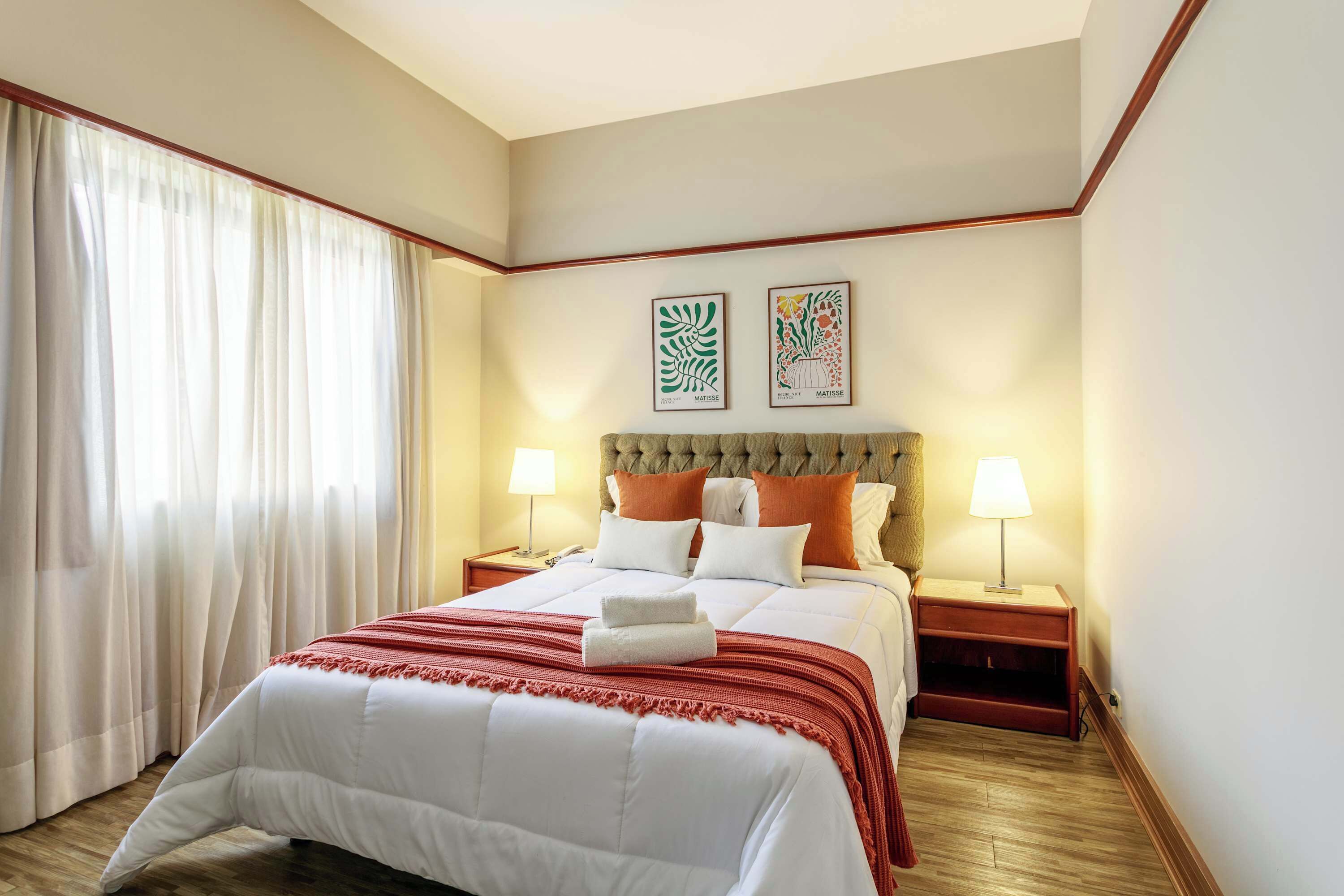 Una habitación de hotel con una cama grande, dos mesitas de noche, un ventanal con cortinas y dos obras de arte colgadas en la pared.