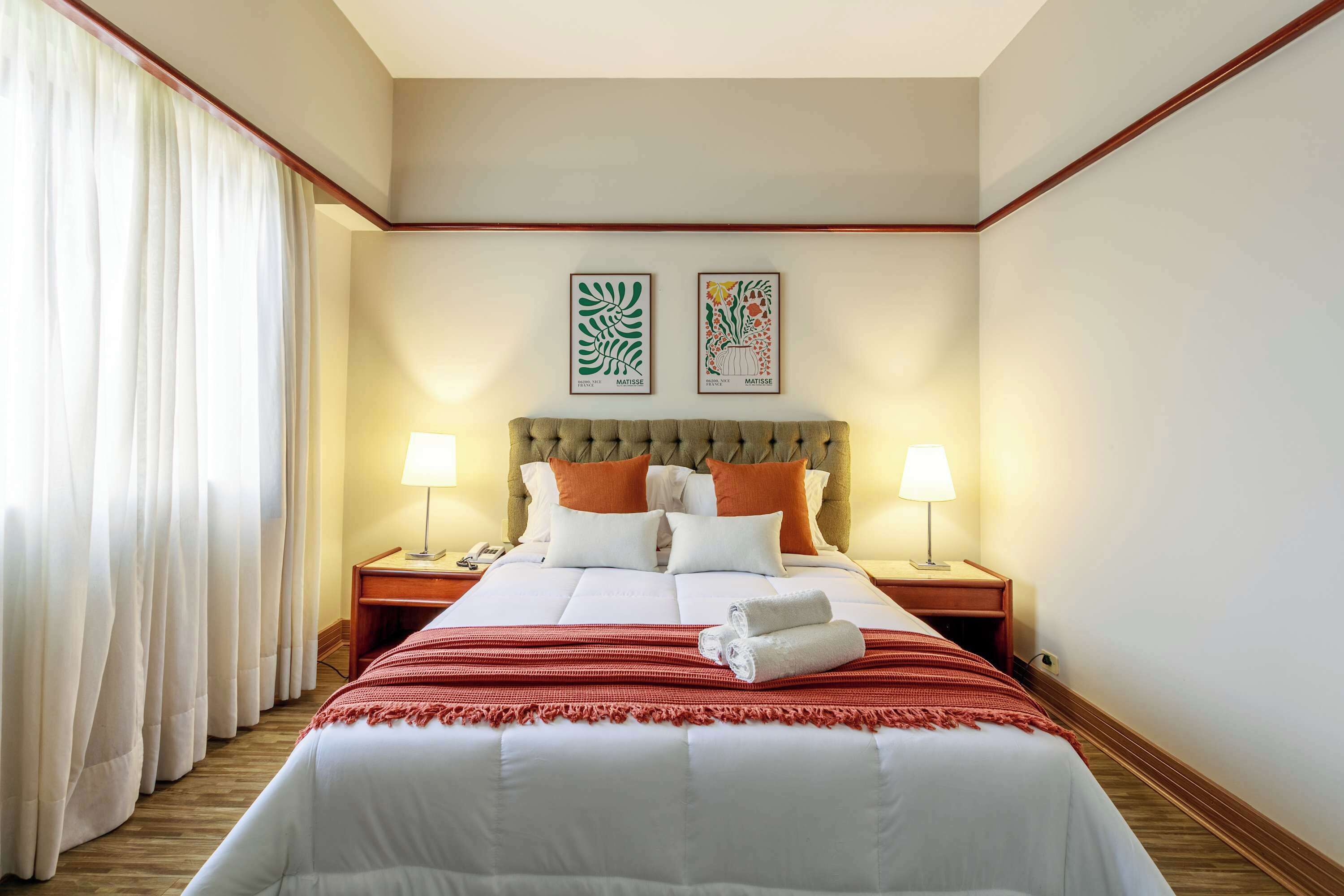 A hotel room with a large bed, two bedside tables with lamps, a headboard, and two framed artworks on the wall.