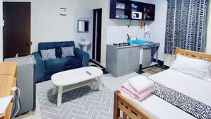A compact hotel room with a bed, a sofa, a small desk, a ceiling fan, a TV, and a bathroom. - Modern Airport Studio Near SGR & Moi Airport (Mombasa)