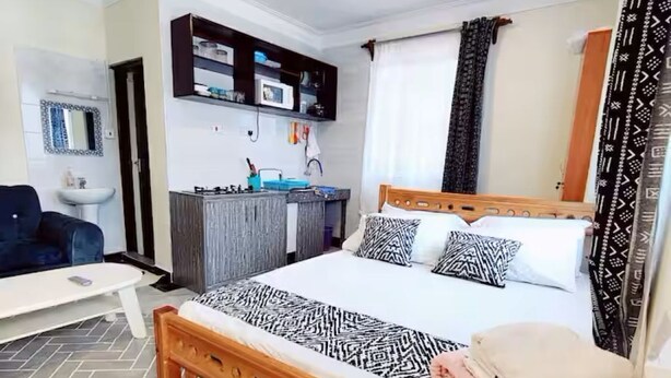 A bedroom with a bed, a ceiling fan, a bedside table, a chair, a small desk, a mirror, a shelf, and a window with curtains. - Modern Airport Studio Near SGR & Moi Airport (Mombasa)