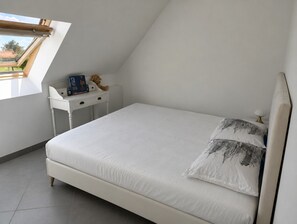 Ein Schlafzimmer mit schrägen Wänden, einem Bett mit weißer Bettwäsche, einem kleinen, weißen Schreibtisch mit Teddybär und Buch sowie einem Fenster mit Blick ins Freie.