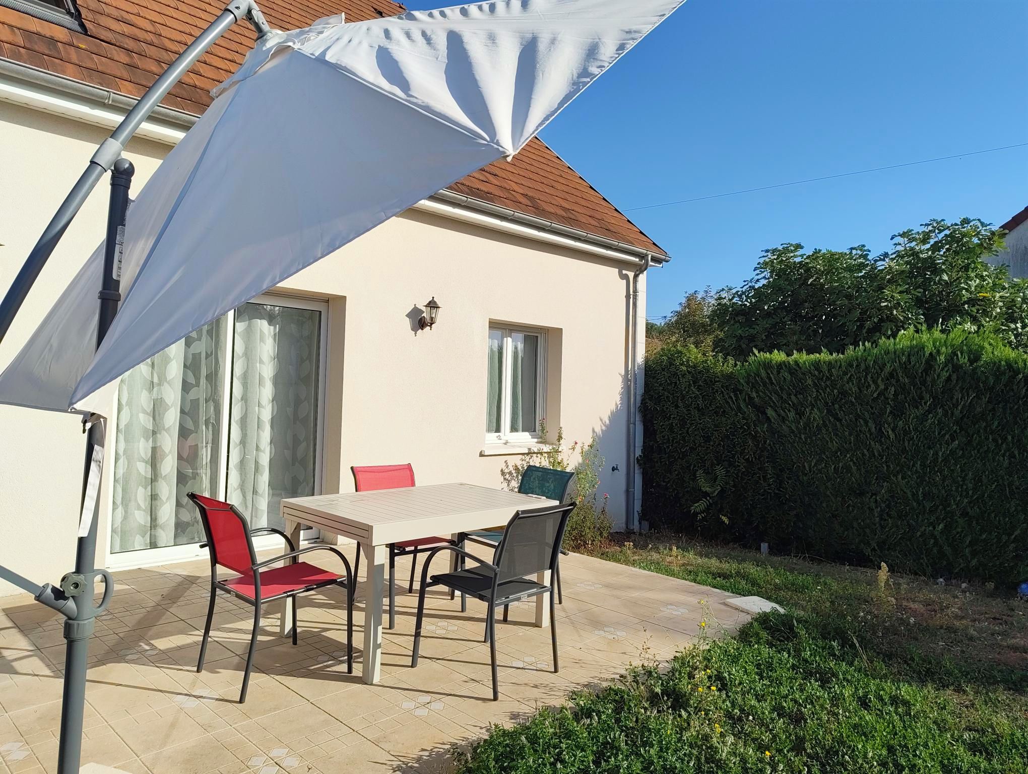 Une terrasse avec une table et des chaises, un parasol blanc et une maison avec un toit rouge.