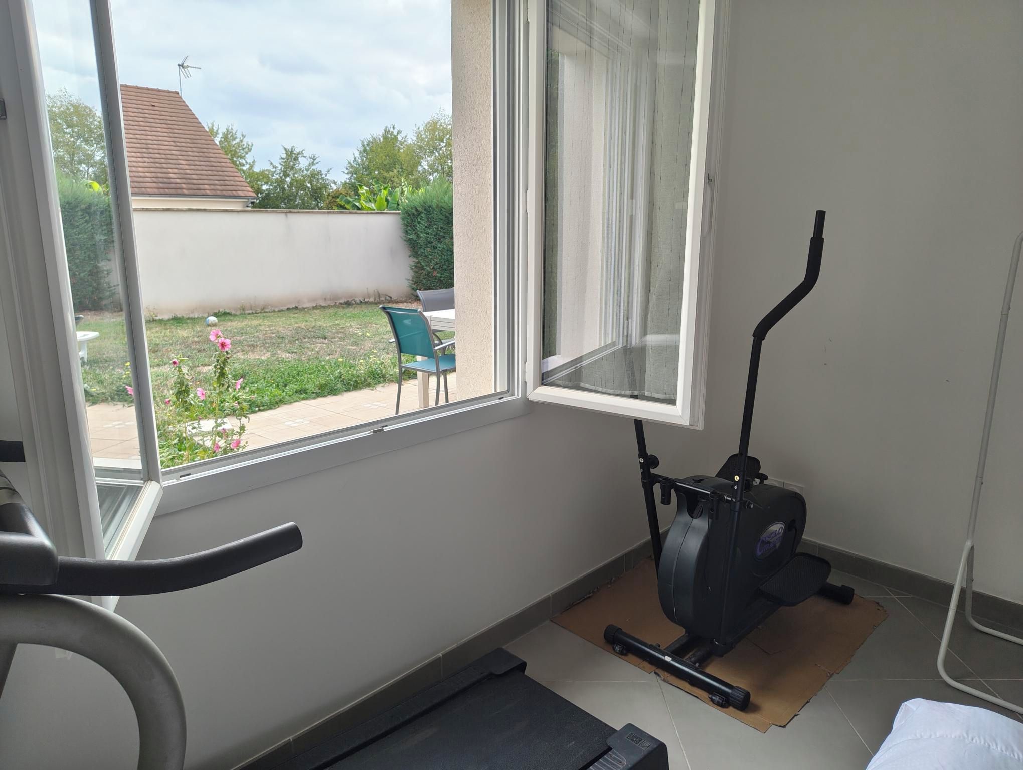A room with a stationary exercise bike, a window with a view of a backyard, and a small table.