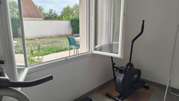 A room with a stationary exercise bike, a window with a view of a backyard, and a small table.