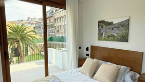 Habitación de hotel con una cama, vistas a edificios y vegetación, un cuadro en la pared y un balcón con una palmera.