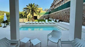 Zona de piscina con sillas reclinables blancas, una palmera y un muro de piedra.