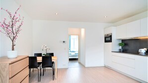 Een moderne keuken met witte kasten, een eethoek met een houten tafel en stoelen, en een zichtbare slaapkamer met een bed en raam.
