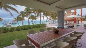 Una terraza de madera con sillas de mimbre, una mesa con frutero y vista al océano y palmeras.