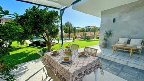 An outdoor patio with a table set for dining, surrounded by greenery and a view of a pool.