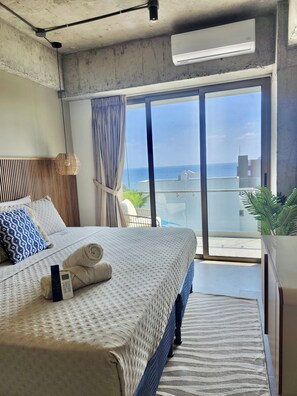 Une chambre avec un lit, une télécommande, une serviette enroulée, une fenêtre donnant sur la mer et un appareil de climatisation.
