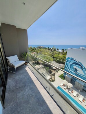 Un balcón con una silla reclinable blanca, barandilla de vidrio, vista al océano y zona de piscina con tumbonas.