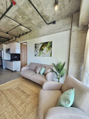 A modern living room with a sofa, a plant, and a kitchen in the background. - Zonte Terrace - 2BR Designer Loft with Surf Views (El Zonte)
