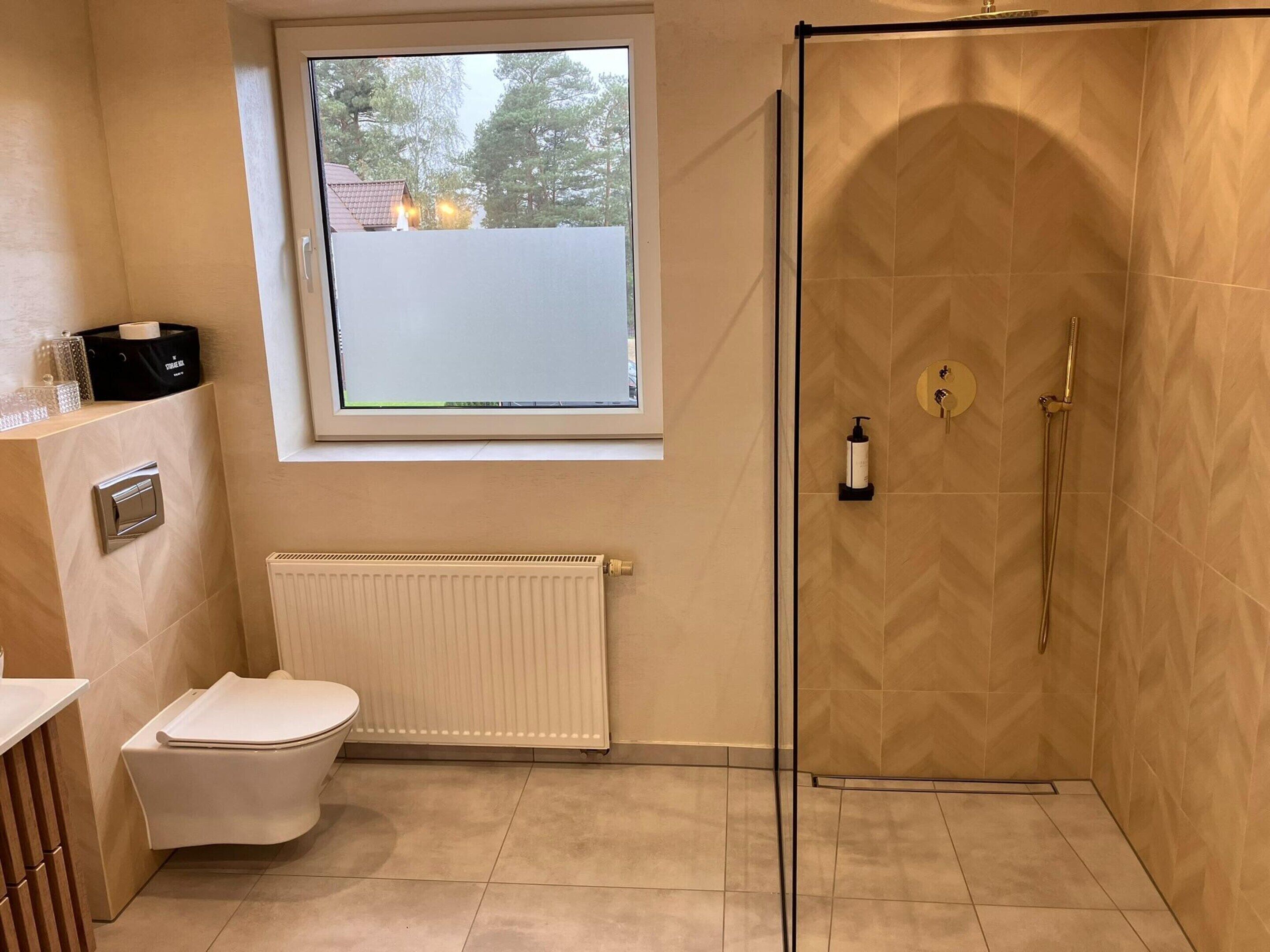 A bathroom with a toilet, a radiator, a shower with a glass door, and a window with a view of trees.