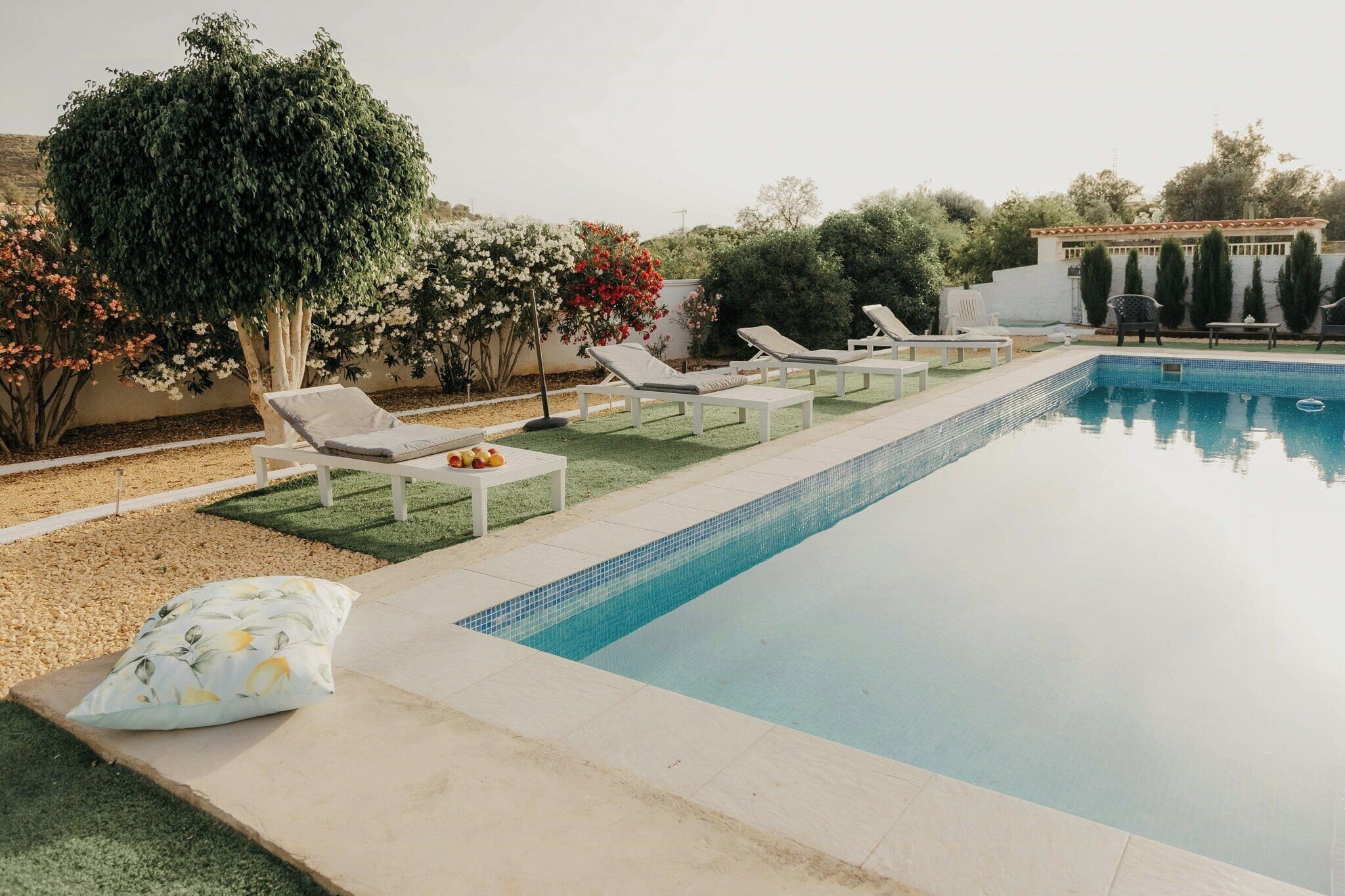 A swimming pool with lounge chairs and a table, surrounded by greenery and a building in the background.