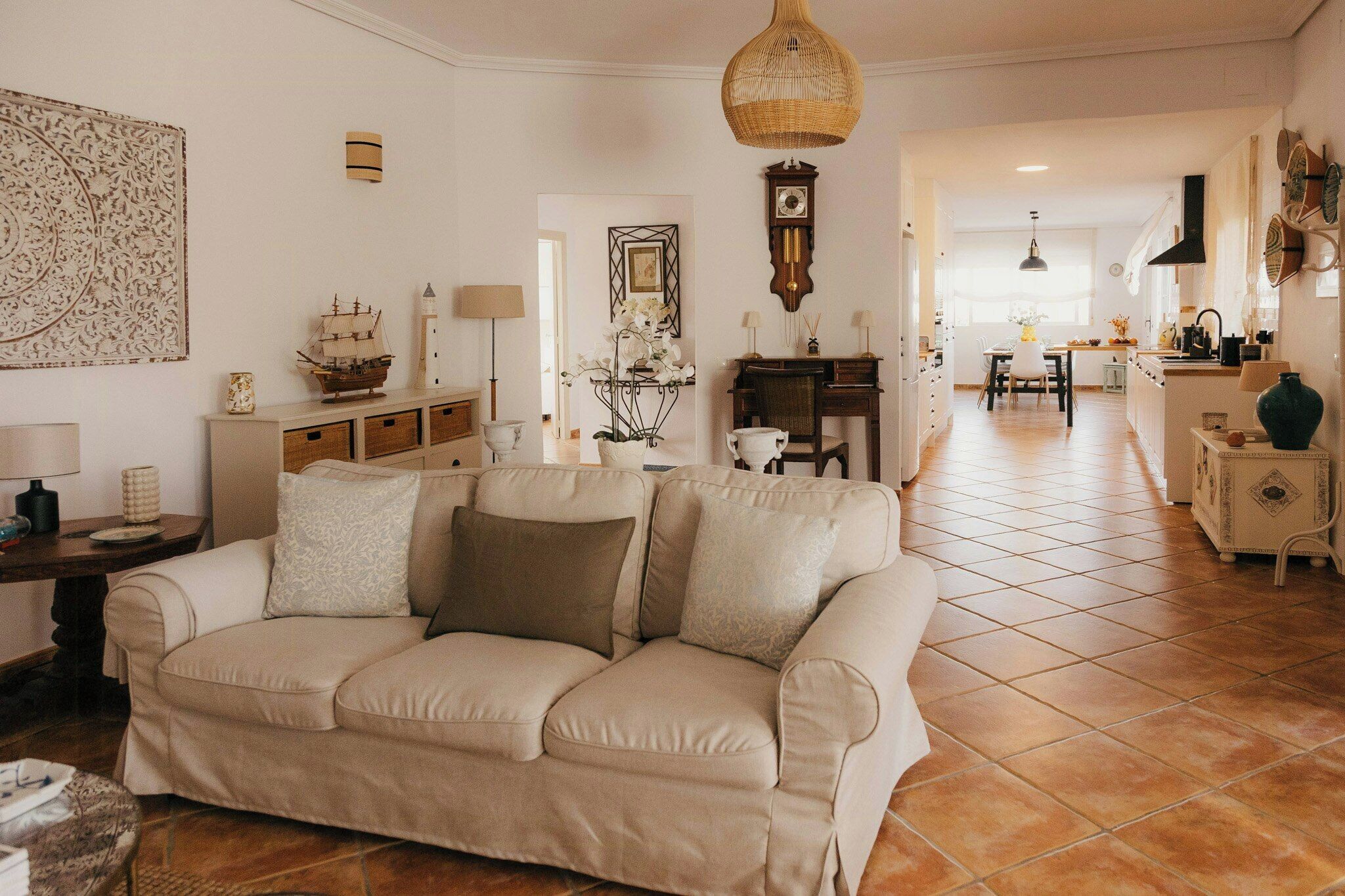 A living room with a beige sofa, a dining area with a table and chairs, a kitchen with a stove and sink, and a hallway leading to another room.