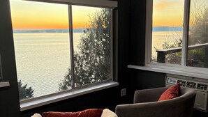 A room with two windows overlooking a body of water during sunset, a wooden floor, and a chair with a cushion.