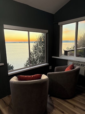 A room with two windows overlooking a body of water during sunset, a wooden floor, and a chair with a cushion. - The Nest - cottage with panoramic views and private deck seating (Burien)