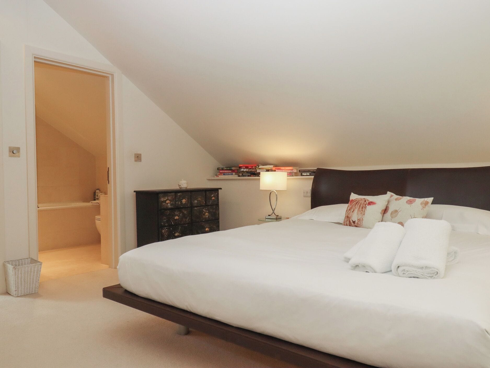 A bedroom with a bed, a nightstand, a dresser, a lamp, and a bathroom visible through an open door.