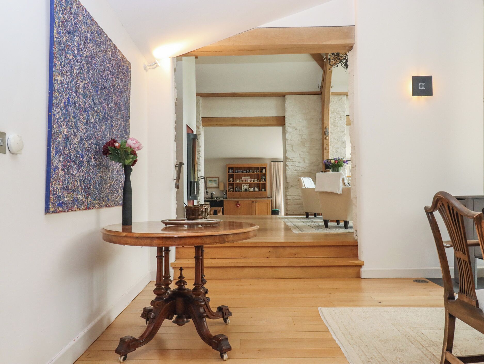 A dining area with a round wooden table, a vase with flowers, and a large abstract painting on the wall.