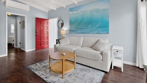 A living room with a beige sofa, a round wooden coffee table, a white nightstand, a red door, and a large wave painting on the wall. - Blue Heron Perch - 5 minute walk to beach, garage (Newport Beach)