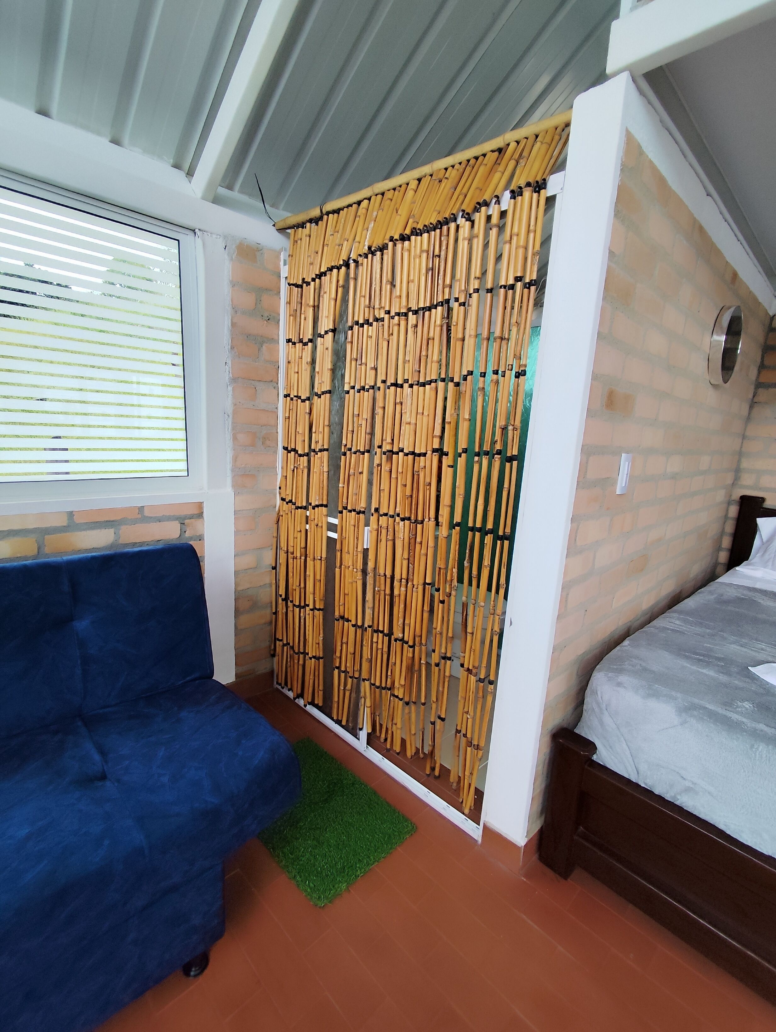 A room with a bed, a blue sofa, a bamboo partition, a window with blinds, and a brick wall.